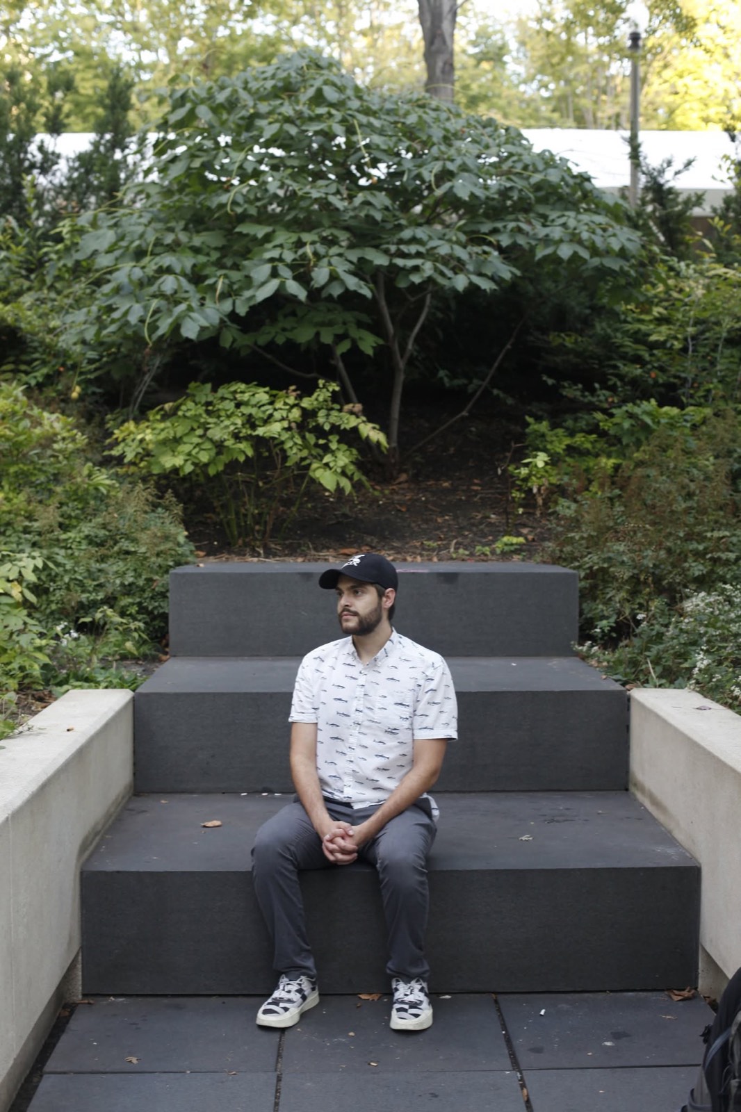 A man sitting on dark stone steps in a leafy garden, hands clasped
